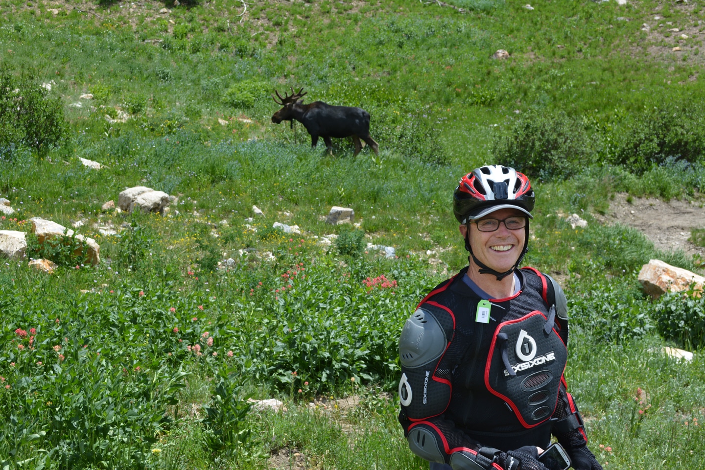 Person in protective gear and helmet smiling with a moose in the background in a grassy, rocky field.